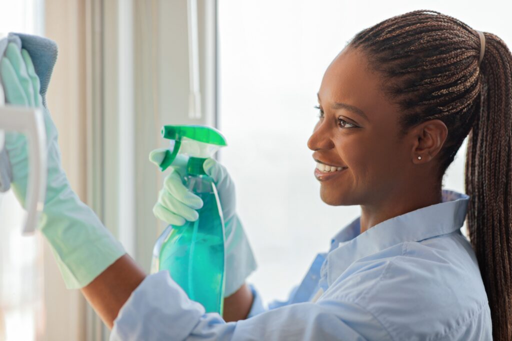 Maid service: Smiling woman cleaning window with spray bottle and cloth. Professional house cleaning services in Washington DC.