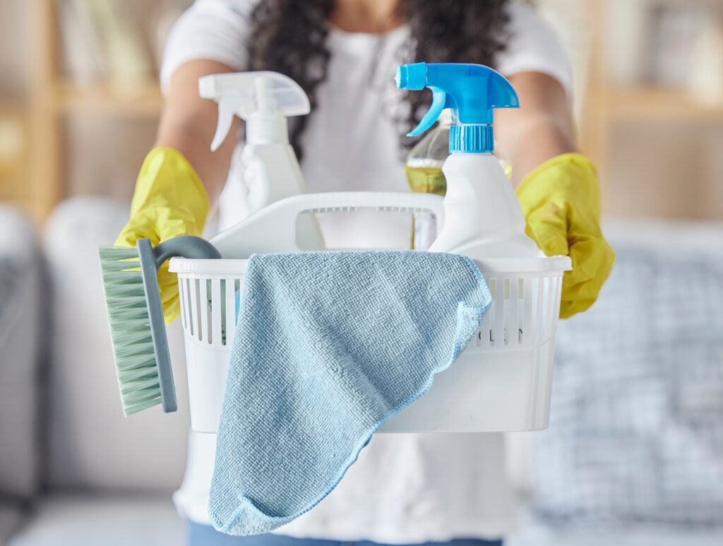 Cleaning supplies in a basket held by a person wearing yellow gloves. Cleaning service tools include spray bottles, brush, and cloth. House cleaning.