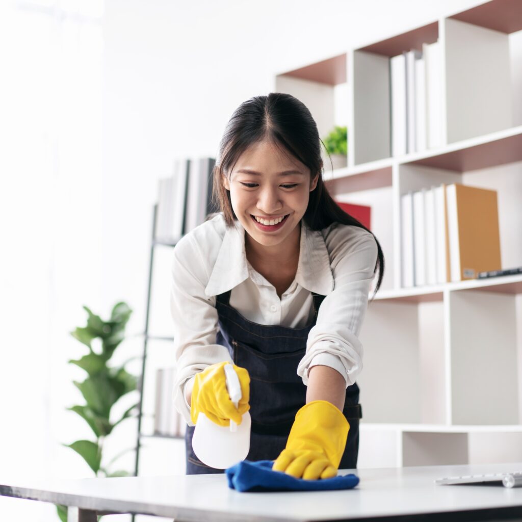 Smiling Asian woman cleaning a desk with spray and cloth, wearing yellow gloves. Office cleaning services in Washington DC.
