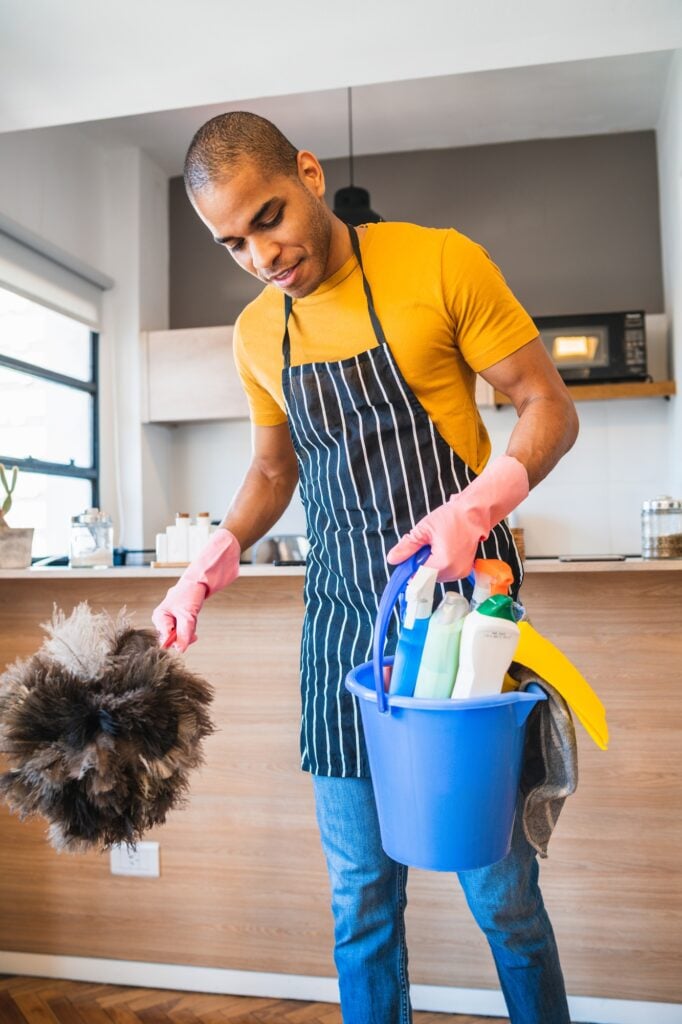Man with duster and bucket of supplies for home cleaning services. Professional cleaner in Washington DC wearing apron and gloves.