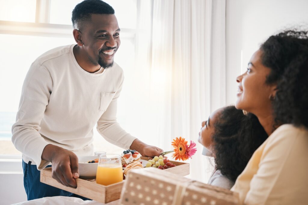 Happy family celebrating with breakfast in bed. Dad brings a tray with food, a flower, and a gift to mom and daughter in Washington DC.