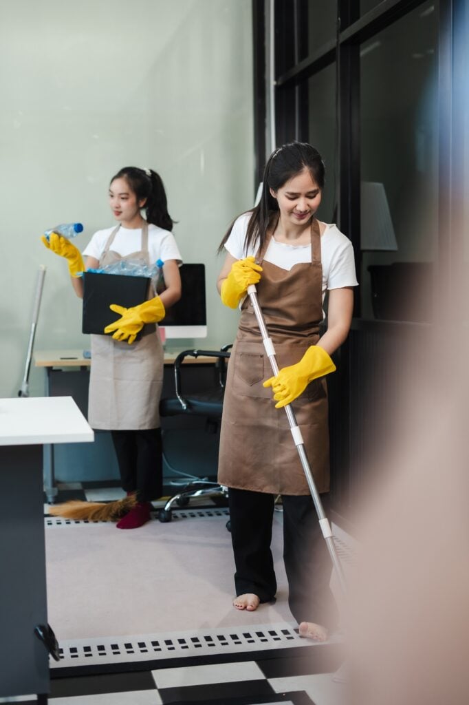 Office cleaning services: two Asian women in aprons and gloves cleaning an office. One is mopping the floor and the other is holding a box of trash.