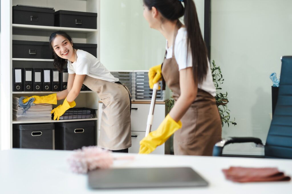 Two women cleaning an office in Washington DC. One is dusting shelves and the other is mopping. They are wearing aprons and yellow gloves. Commercial Cleaning.
