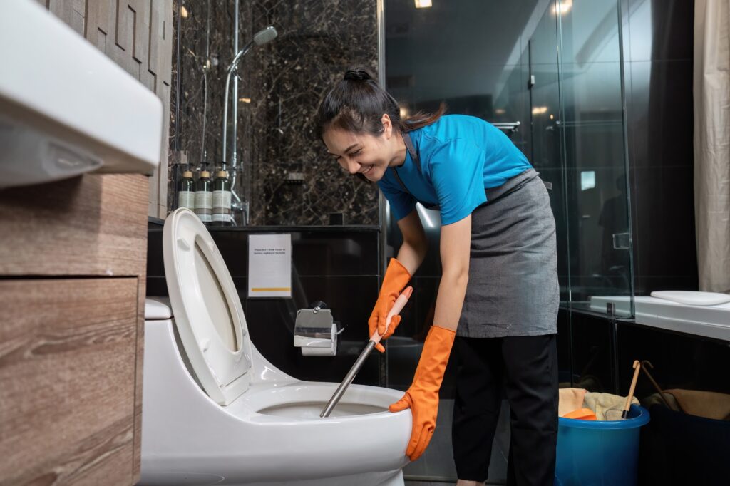 Bathroom cleaning services: Woman cleaning a toilet with a brush, wearing gloves and smiling. Professional house cleaning services in Washington DC.