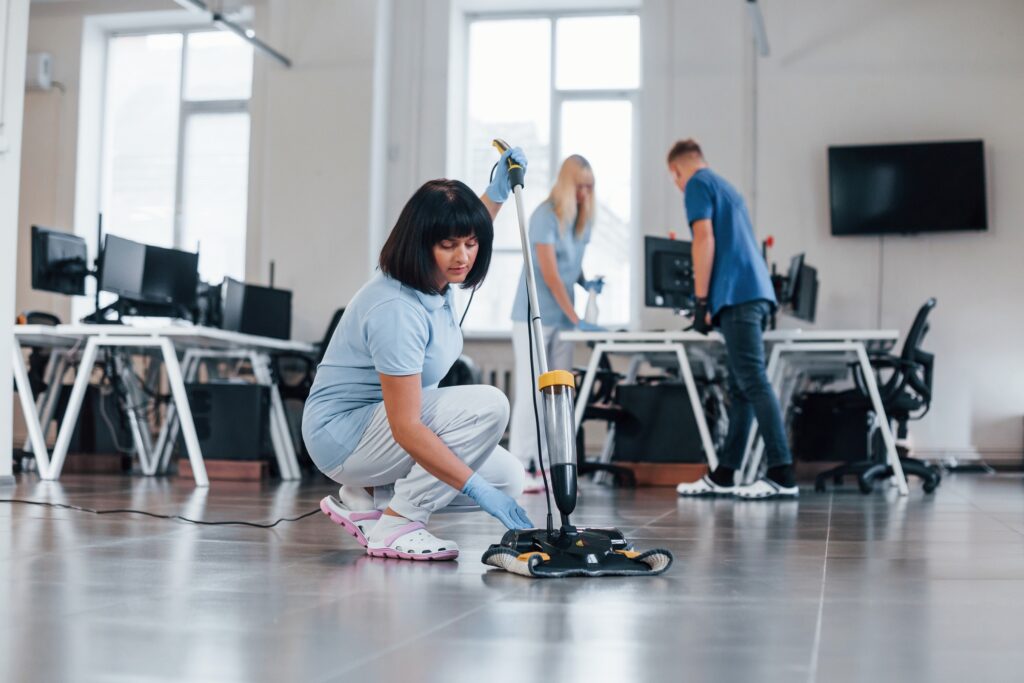 Office cleaning services team. Woman using floor cleaning machine in Washington DC, while colleagues clean desks. Commercial cleaners.