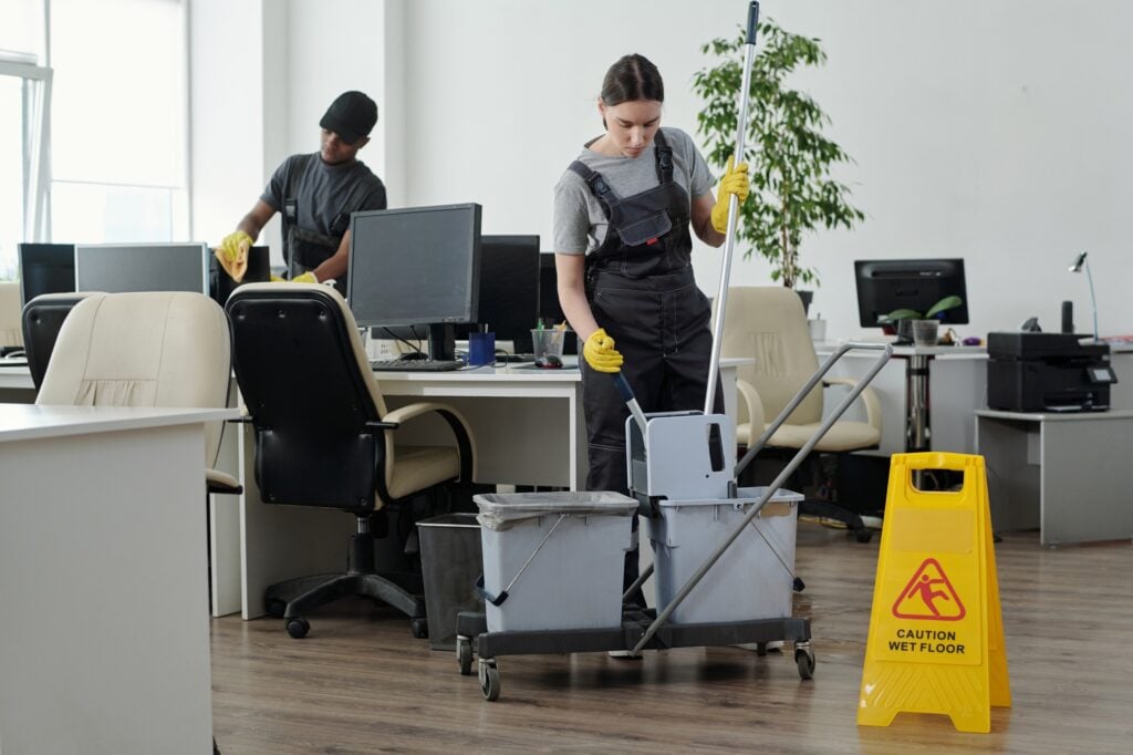 Office cleaning services: Two cleaners mopping and wiping down desks in an office with a wet floor sign. Commercial cleaning services in Washington DC.