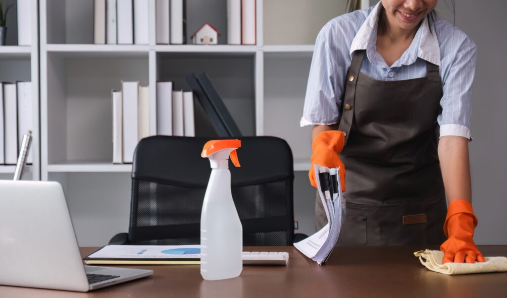 Office cleaning services: Smiling woman dusting desk with laptop, spray bottle, and documents. Professional cleaner in Washington DC wiping surfaces.