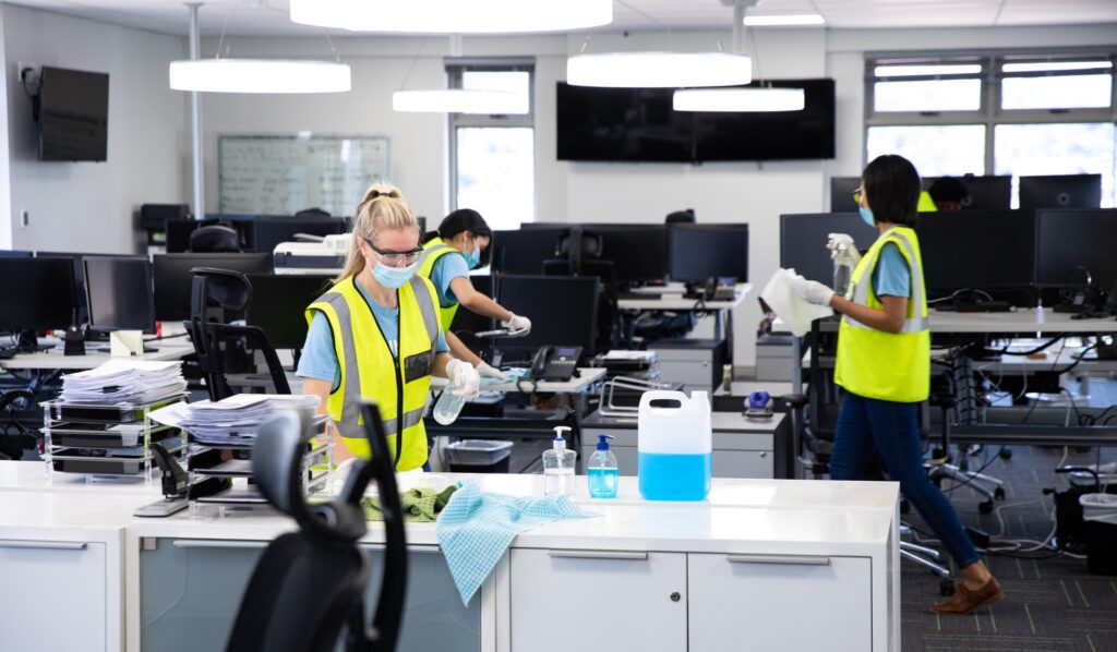 Office cleaning services: Cleaners in face masks disinfecting desks with spray bottles and wipes in a modern office space, Washington DC.