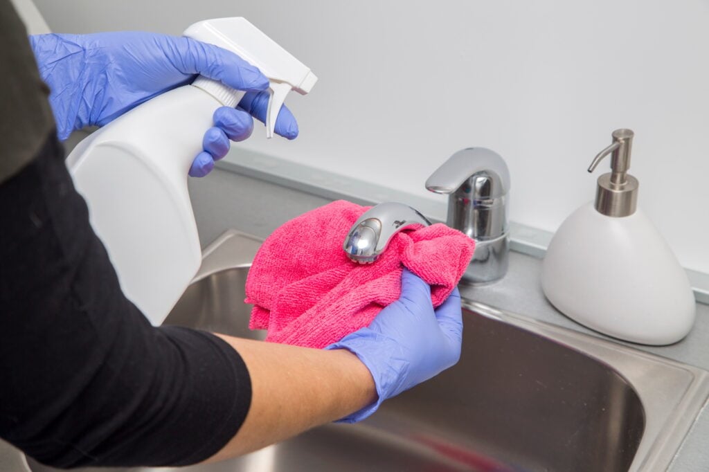 Bathroom cleaning service: gloved hands wipe a faucet with a pink cloth and spray bottle. Professional home cleaning in Washington DC.