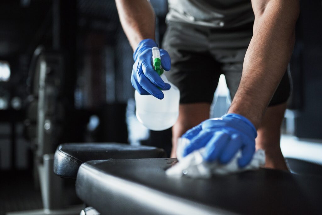 Gym equipment cleaning service. A person in blue gloves wipes down a bench with a spray bottle and cloth. Fitness center hygiene.