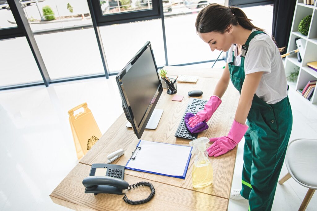 Office cleaning services: Woman in pink gloves wipes keyboard with a cloth. Desk has a phone, monitor, and spray bottle. Wet floor sign is visible.