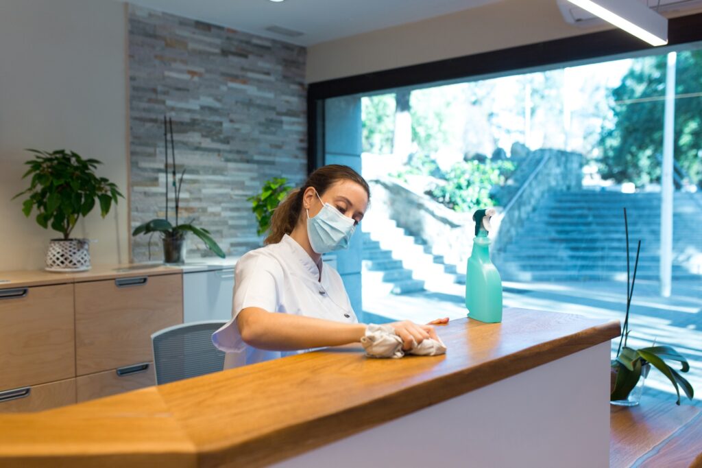 Woman wearing a mask cleaning the reception desk with a cloth and a spray bottle. Professional office cleaning services in Washington DC.