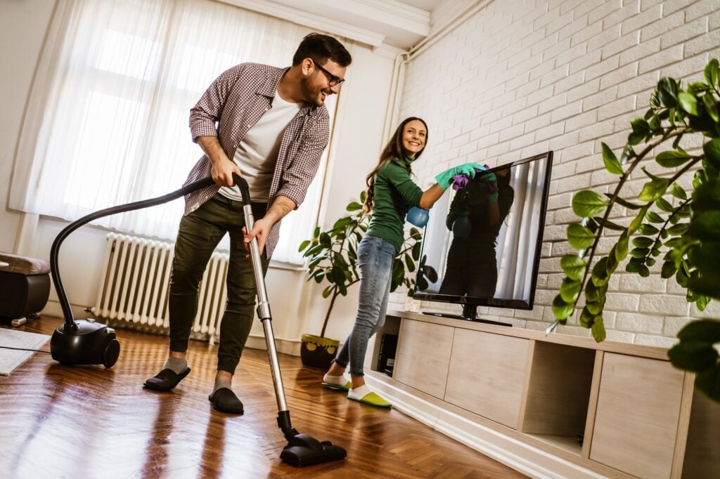 Couple cleaning home. Man vacuums hardwood floor while woman cleans TV screen. Residential cleaning services in Washington DC.