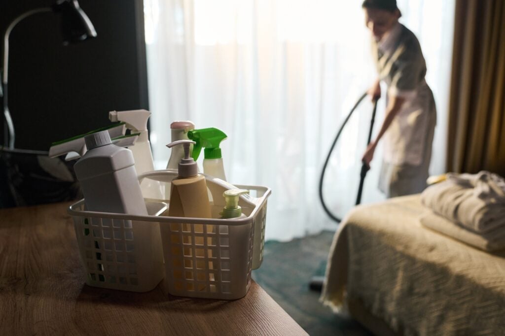 Maid service in Washington DC vacuuming a hotel room. Cleaning supplies in a basket on a wooden table. Professional housekeeping services.