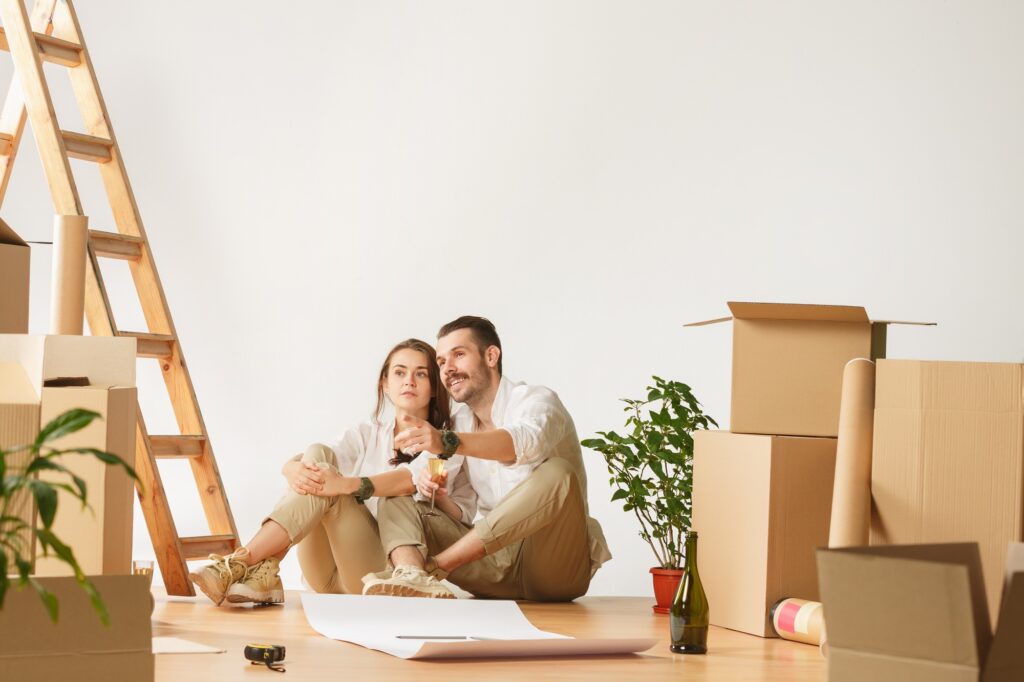 Couple plans home renovation in Washington DC. Happy homeowners celebrate with champagne amidst moving boxes and a ladder, ready for their new space.